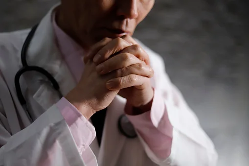 Hands clasped in prayer, a physician in a white coat with a stethoscope
